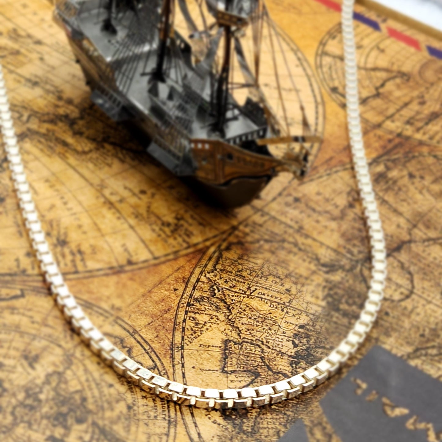 Cadena tipo caja veneciana de plata italiana 925, colocada sobre un mapa antiguo junto a un barco pequeño de metal, evocando la aventura, la exploración y la sofisticación de la marca Nosso Galeón.