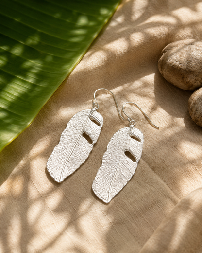  Sterling silver banana leaf dangle earrings resting on a fresh green banana leaf, handmade in Colombia.